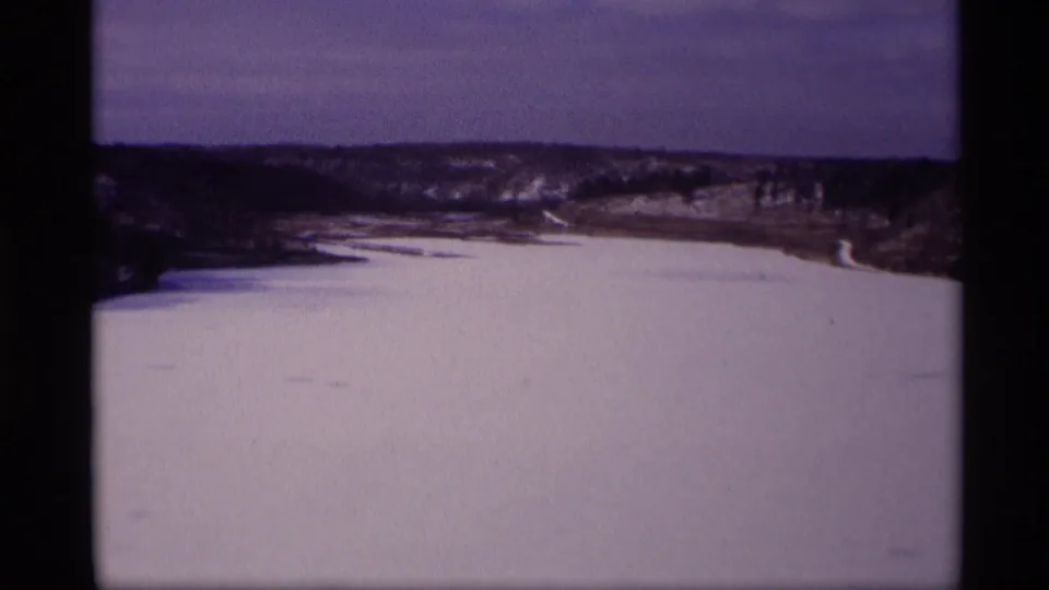 1969 Wisconsin 8mm frame of a broad frozen water surface, pale snowbanks, and wooded hills seen from an elevated home-movie viewpoint.