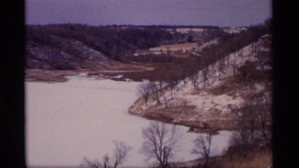 1969 Wisconsin 8mm frame showing a frozen bend bordered by bare trees, patchy snow, exposed thawing ground, and low wooded slopes.