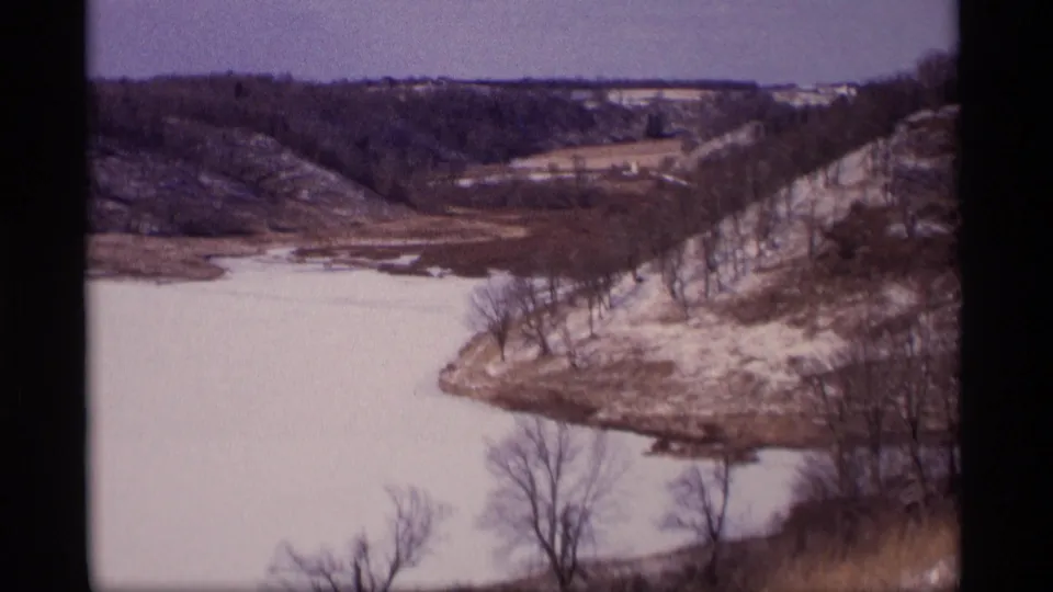 1969 Wisconsin 8mm frame of a frozen shoreline bend, leafless trees, snow patches, and distant wooded hills in cold weather.