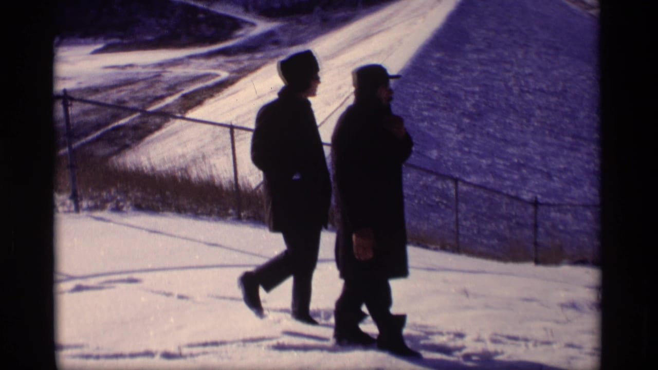 Two men in dark winter coats walk across snow beside a levee railing.