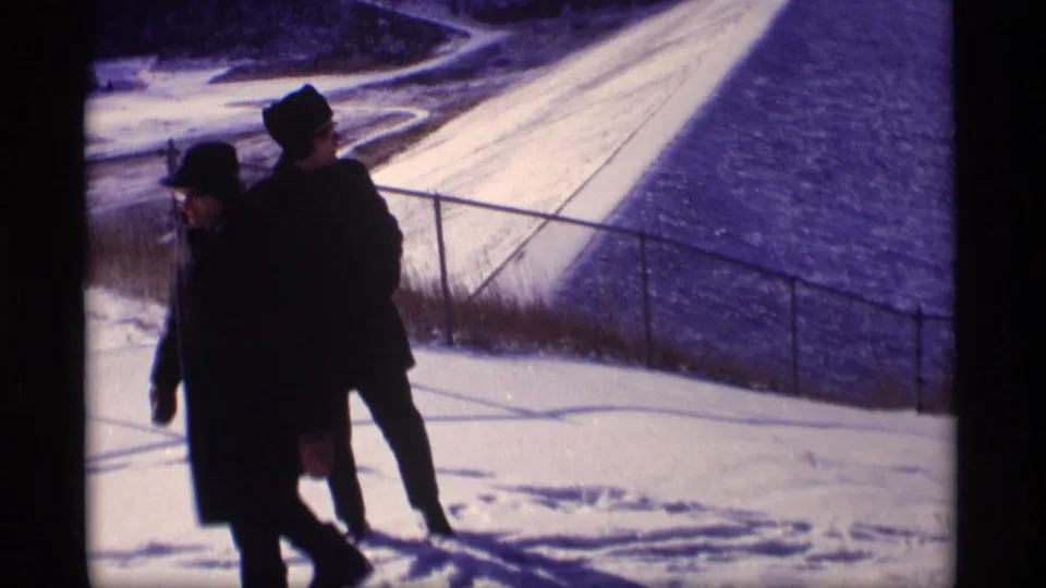 Two men in dark coats pass close to the camera as the snowy levee slope and railing fill the background.