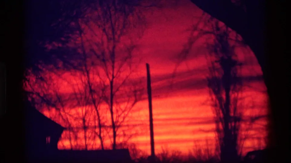 1969 Wisconsin 8mm frame showing a bright orange sunset with dark bare trees, a roofline, and a vertical utility pole.