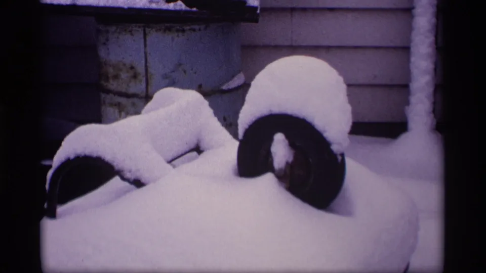 A 1969 Wisconsin winter frame showing fresh snow over a low yard object near house siding, with dark curved tire-like forms partly exposed.