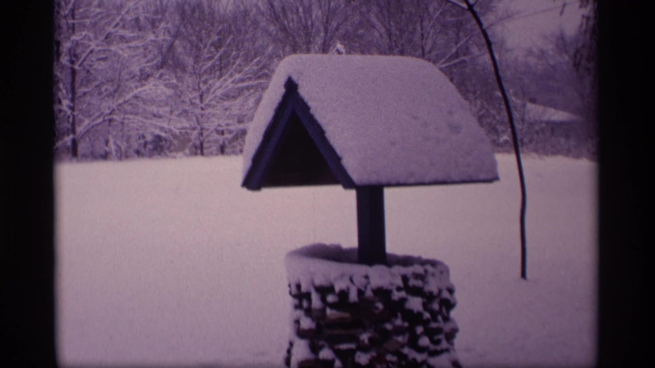 1969 Wisconsin 8mm frame of a snow-covered roofed wishing well set against trees and an open winter landscape.