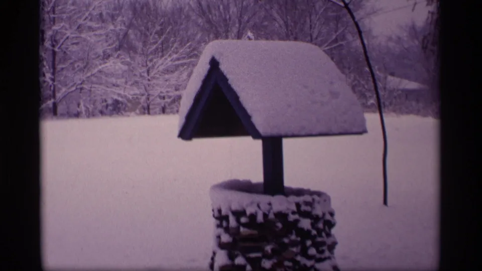 1969 Wisconsin 8mm frame of a snow-covered roofed wishing well set against trees and an open winter landscape.