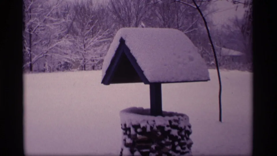 1969 Wisconsin 8mm frame showing a roofed wishing well blanketed in snow with winter trees in the background.
