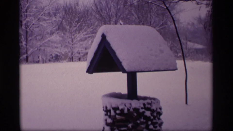 1969 Wisconsin 8mm frame of a small yard wishing well under heavy snow, with wooded winter background.