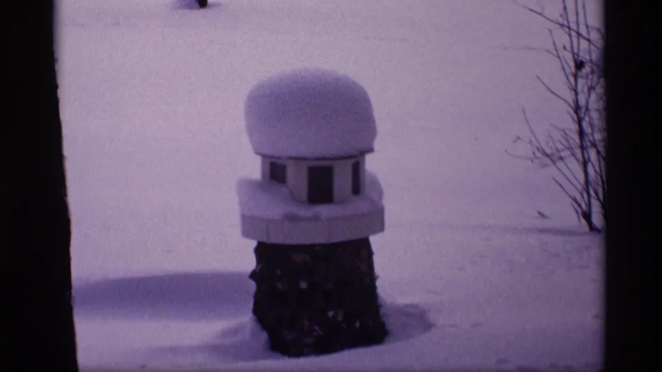 1969 Wisconsin winter frame with a rounded snow cap on a small birdhouse or feeder set against a snowy yard.