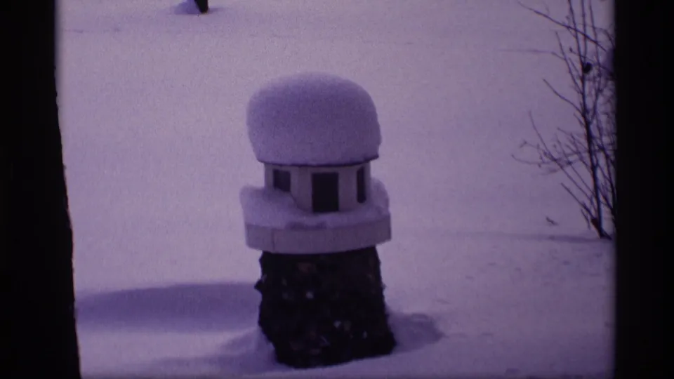 1969 Wisconsin 8mm frame showing a small birdhouse or feeder with heavy snow, framed by dark trunks and bare winter brush.