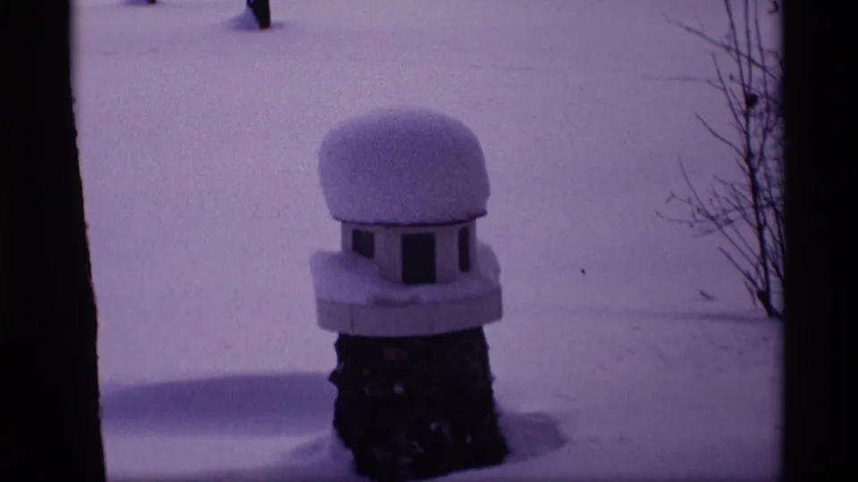 1969 Wisconsin winter frame focused on a snow-capped birdhouse or feeder near a dark tree trunk in a quiet snowy yard.