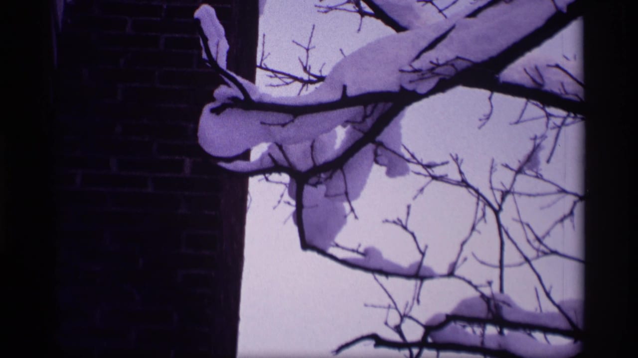 Snow-covered branches beside a dark brick wall against a pale winter sky.