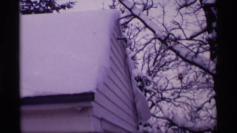 1969 Wisconsin home-movie frame showing heavy snow on a white-sided house and leafless tree branches against a pale sky.
