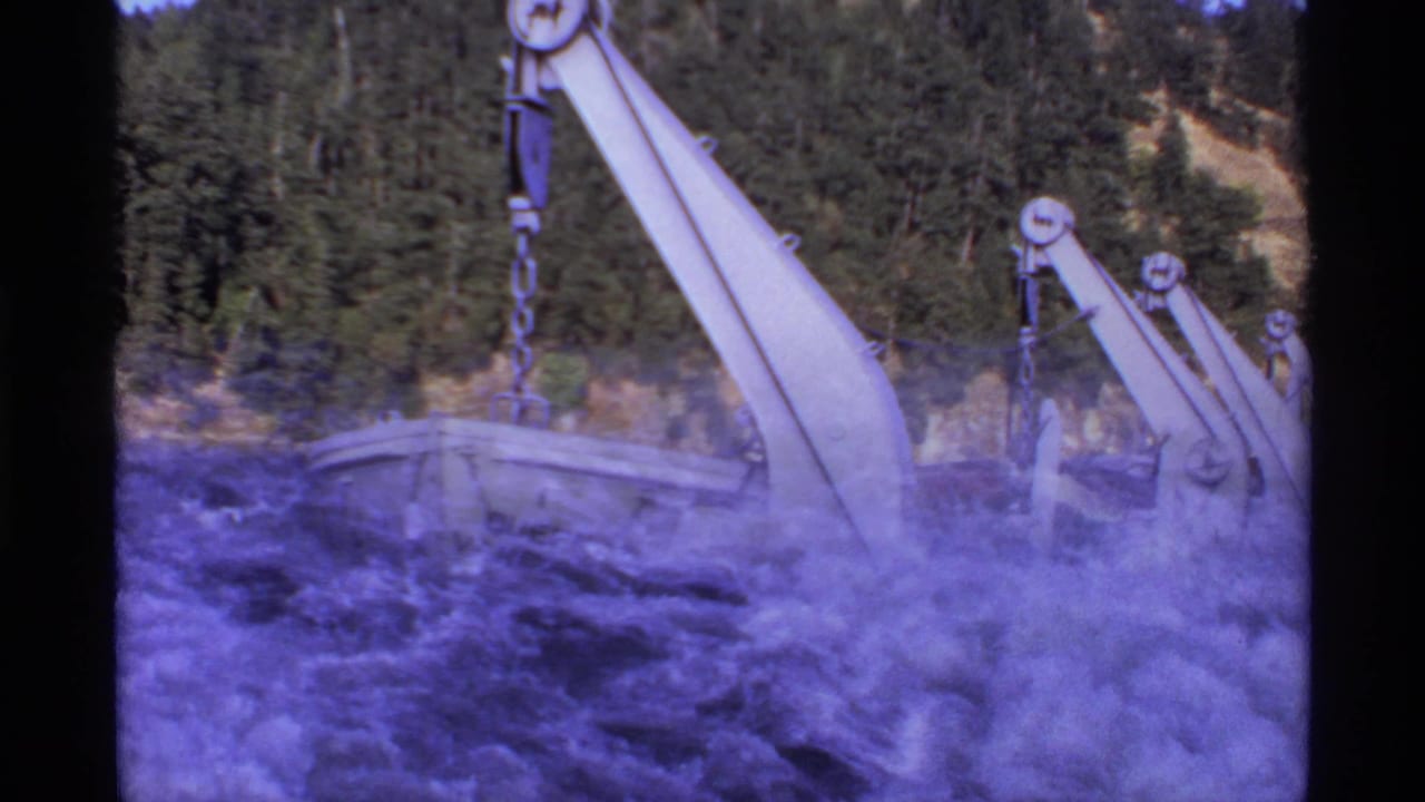 Lifeboats and davit arms above turbulent water with forested shoreline behind.