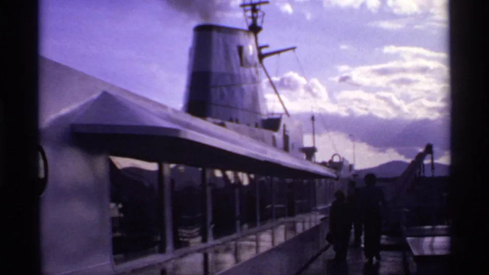 1974 Canadian home-movie frame showing a ship funnel smoking above a covered deck while passengers stand or walk nearby.