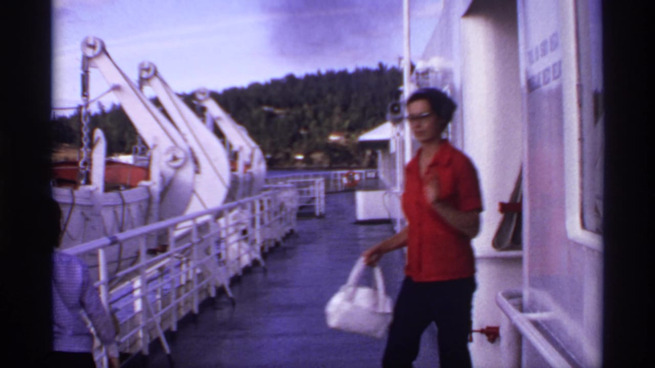 1974 Canada home-movie frame of a woman walking on a ferry deck with white railings, davit arms, forested shoreline, and cloudy sky.
