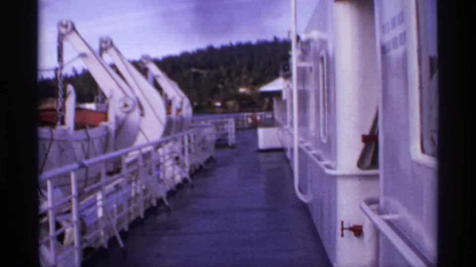 1974 Canada ferry deck after the passerby leaves frame, showing white railings, davit arms, ship wall, and a forested shore.