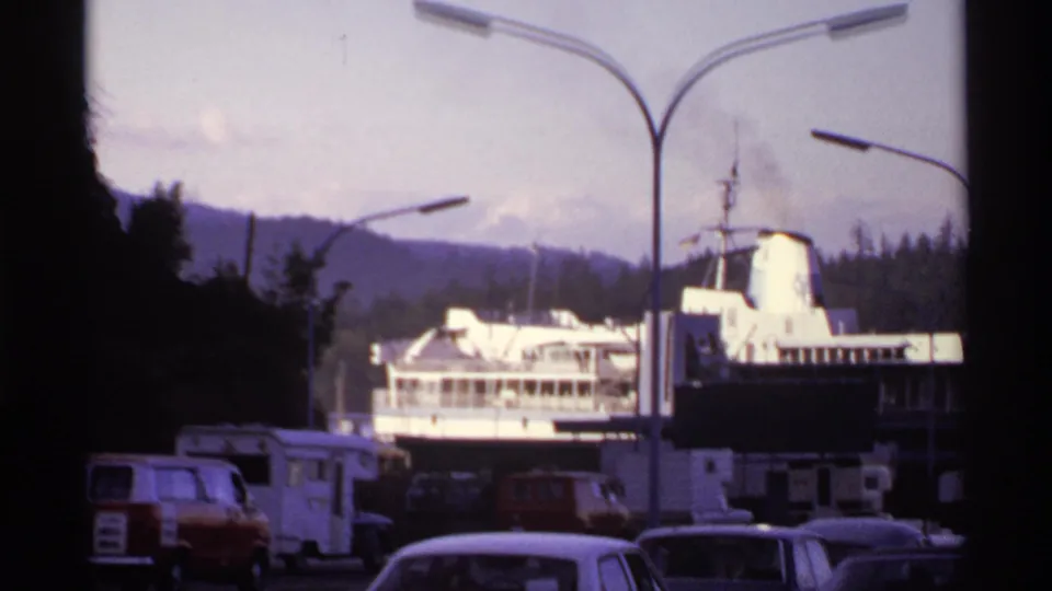 1974 Canada terminal view with a docked ferry, parked vintage cars and RVs, tall lamp posts, mountains or wooded hills, and cloudy sky.