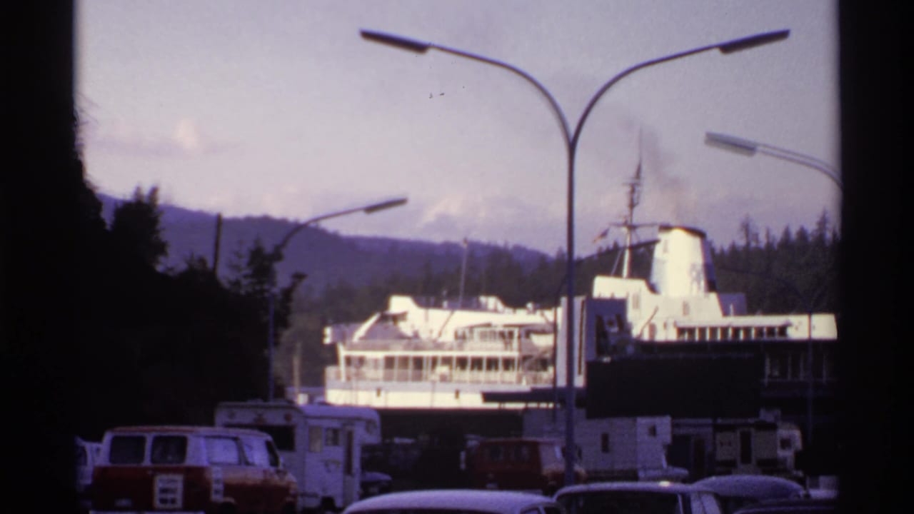 Docked ferry beyond parked vans and campers near tall streetlights and wooded hills.