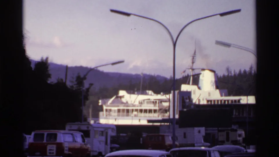 1974 Canadian 8mm frame showing a docked ferry seen past parked vans, campers, streetlights, trees, and distant hills.