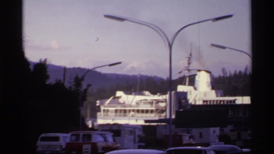 1974 Canadian waterfront view with a docked ferry framed by modern streetlights, parked vehicles, trees, and distant hills.