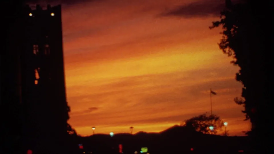 1974 Canadian 8mm sunset frame with a tower silhouette, dark trees, distant hills, and small lights under orange sky.