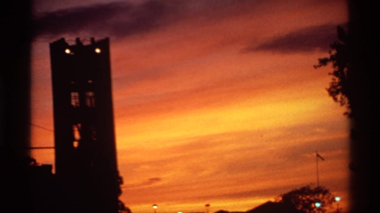Dark tower silhouette against a vivid orange sunset sky with faint city lights below.