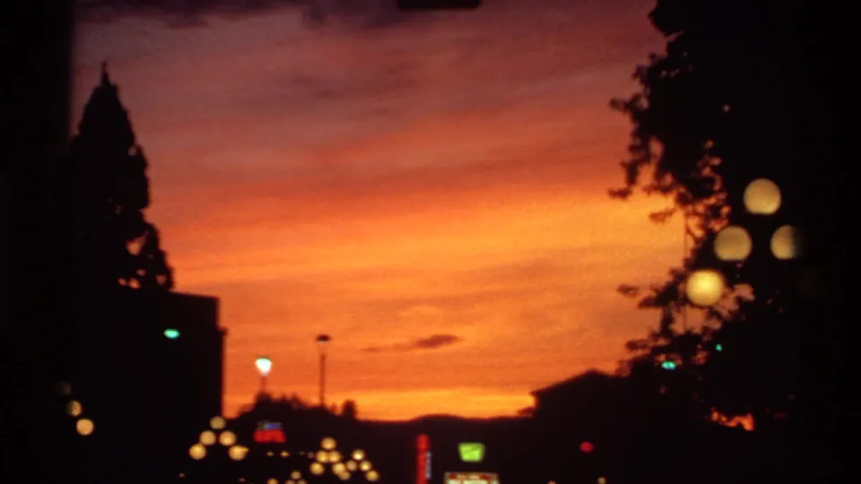 1974 Canadian 8mm frame looking past dark trees and buildings toward glowing streetlights beneath a vivid orange sunset sky.
