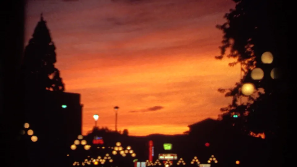 1974 Canadian home-movie frame with streetlights, tree silhouettes, low rooftops, and an orange dusk sky fading toward the dark urban horizon.