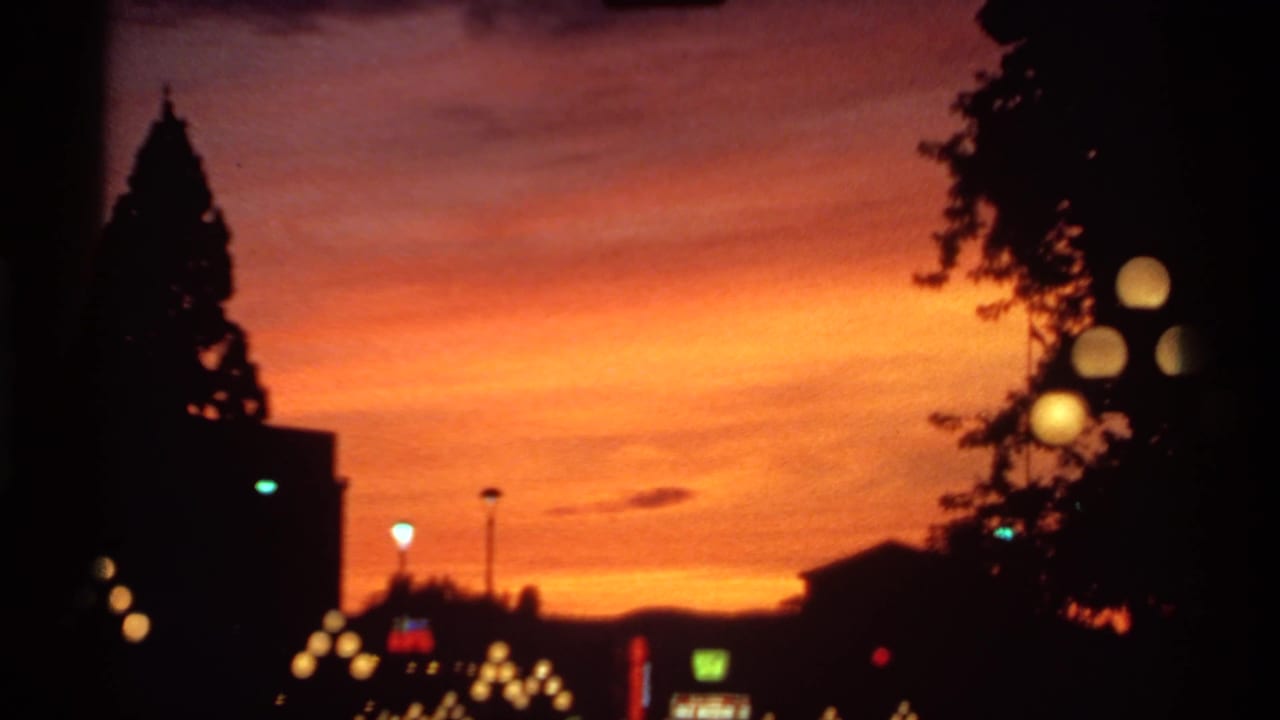1974 Canadian 8mm frame showing a vivid orange dusk sky, silhouetted trees and buildings, and soft glowing streetlights.