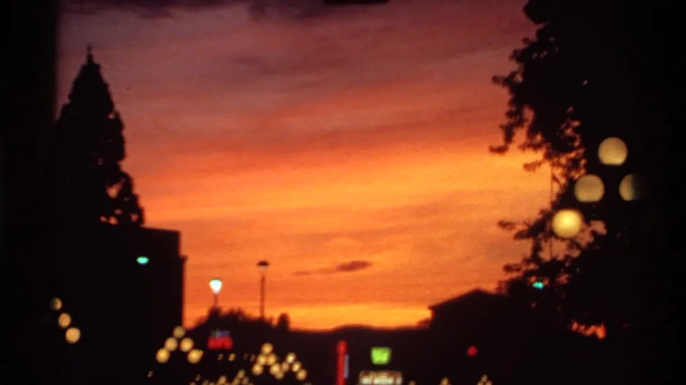 1974 Canadian 8mm frame showing a vivid orange dusk sky, silhouetted trees and buildings, and soft glowing streetlights.
