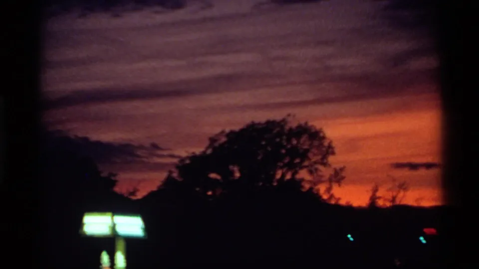 1974 Canadian 8mm frame with a purple-orange evening sky, dark trees, low horizon forms, and an overexposed illuminated sign.