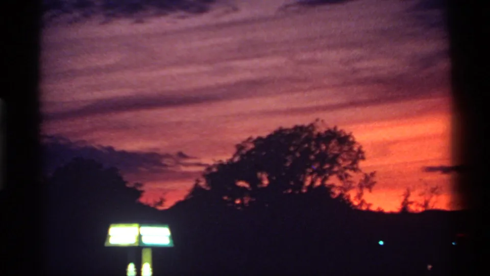 1974 Canadian 8mm frame showing layered purple cloud bands, a red-orange horizon, silhouetted trees, and a glowing rectangular sign.