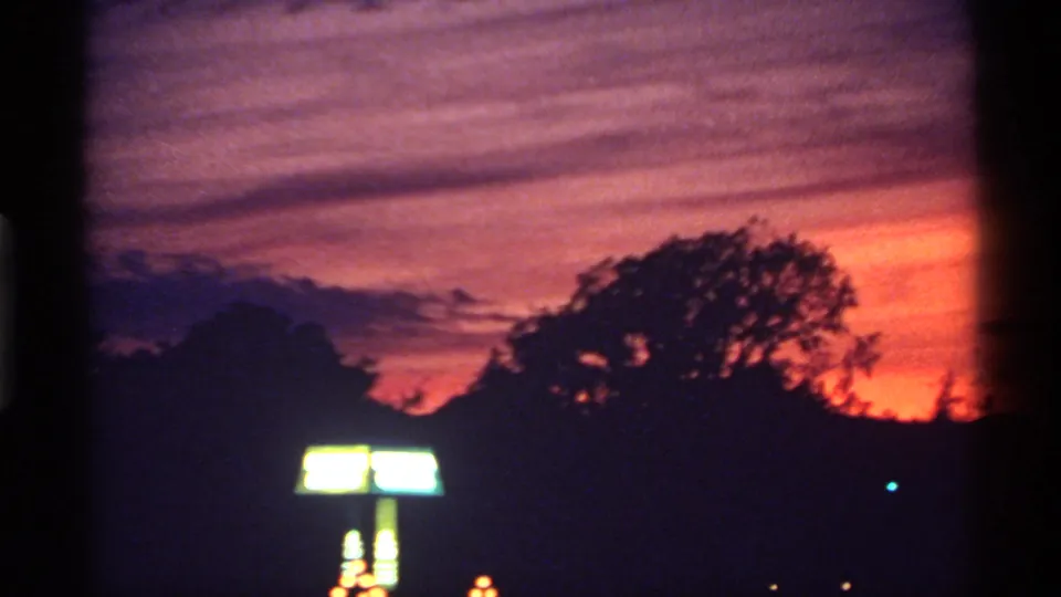 1974 Canadian 8mm frame capturing a dark town or roadside edge beneath orange-purple dusk, with silhouetted trees and a bright sign.