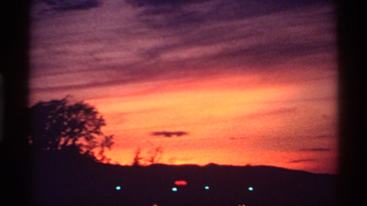 Orange and purple sunset over dark trees, low hills, and small distant lights.