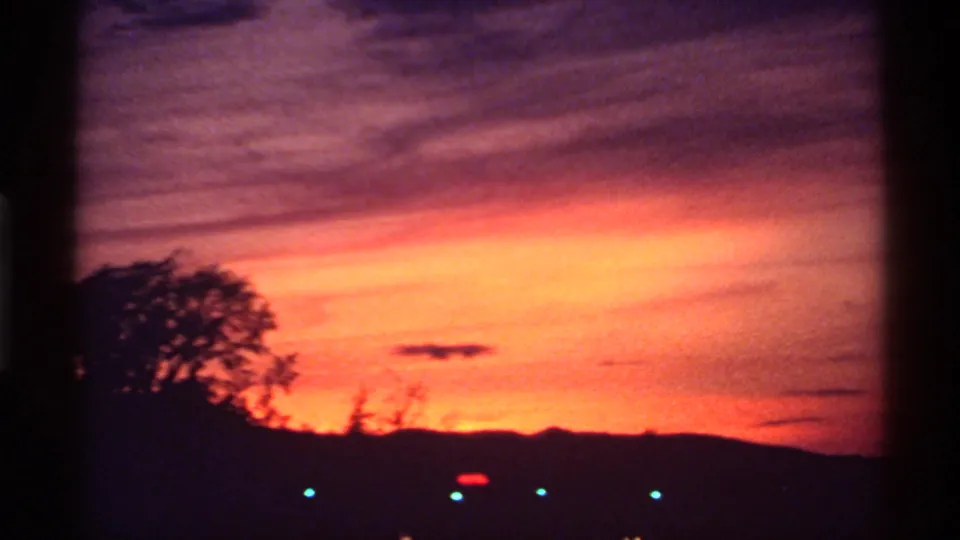 1974 Canada 8mm frame showing a vivid sunset over silhouetted trees, low hills, and faint city lights below.