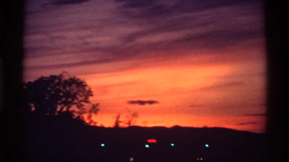 1974 Canada 8mm frame with orange-purple clouds over a dark tree silhouette, low ridge, and small lights.