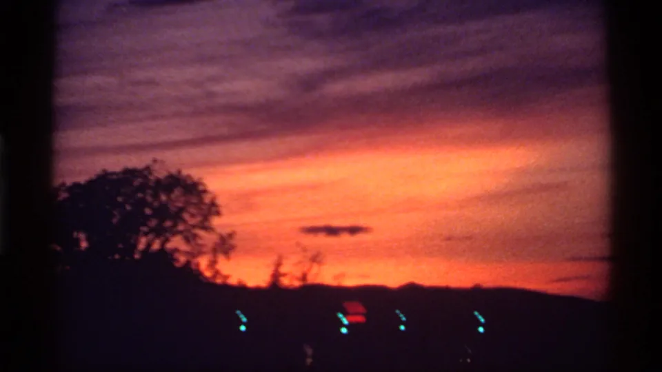 1974 Canada 8mm frame emphasizing saturated sunset color over silhouetted trees, hills, and a few distant lights.