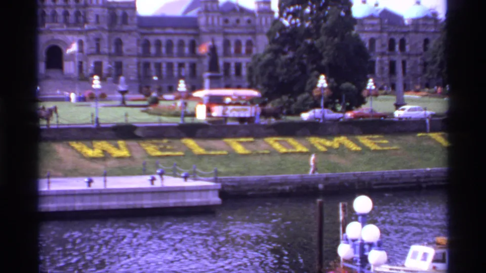 1974 Canada 8mm frame showing a waterfront welcome display, lampposts, cars, pedestrians, and a large civic building beyond.