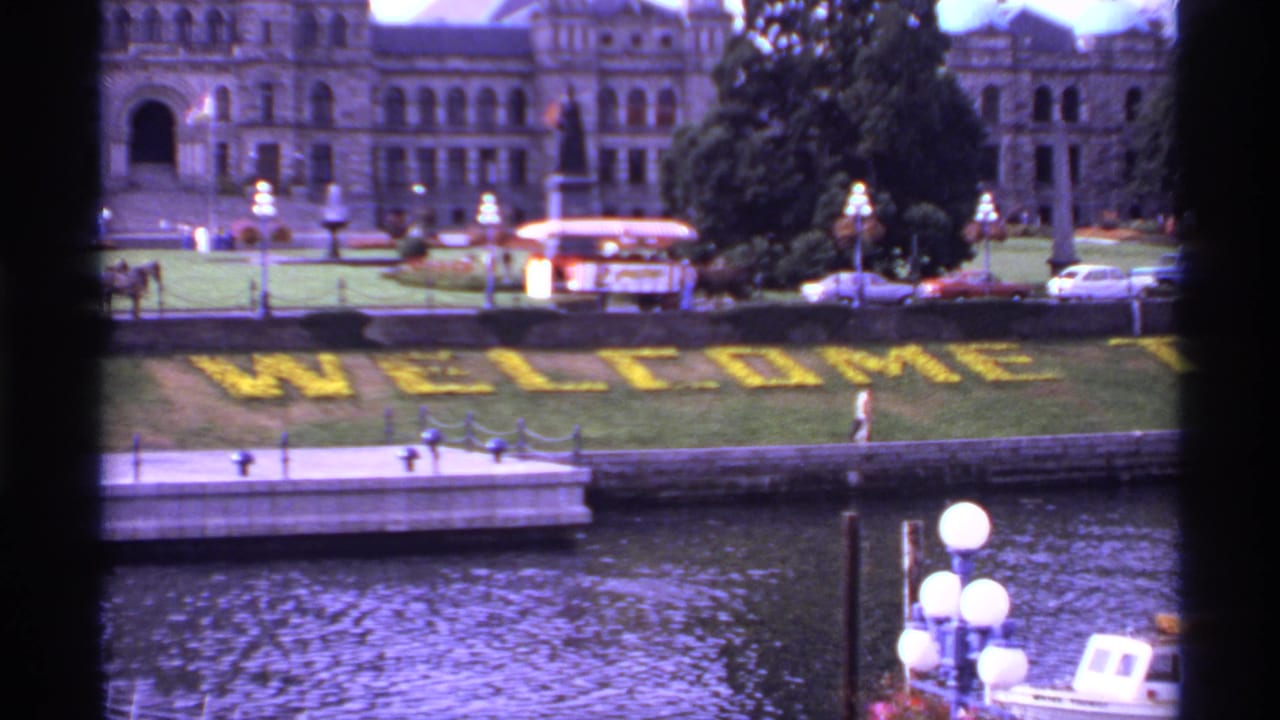 1974 8mm view of Victoria, Canada, with yellow floral welcome lettering beside water, classic cars, lamps, and a large historic-looking building.