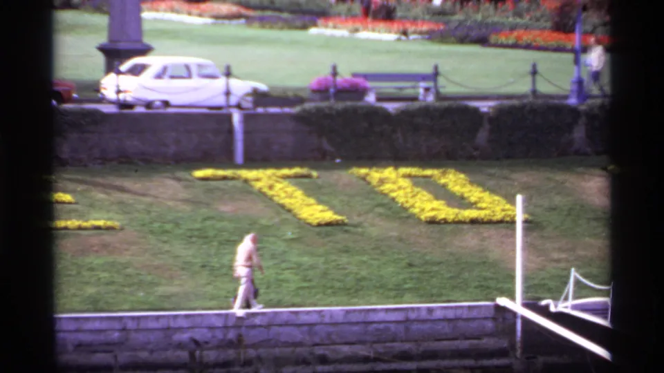 Victoria waterfront lawn with yellow flower lettering, a distant walker, stone wall, white car, bench, and garden plantings in 1974 home-movie footage.