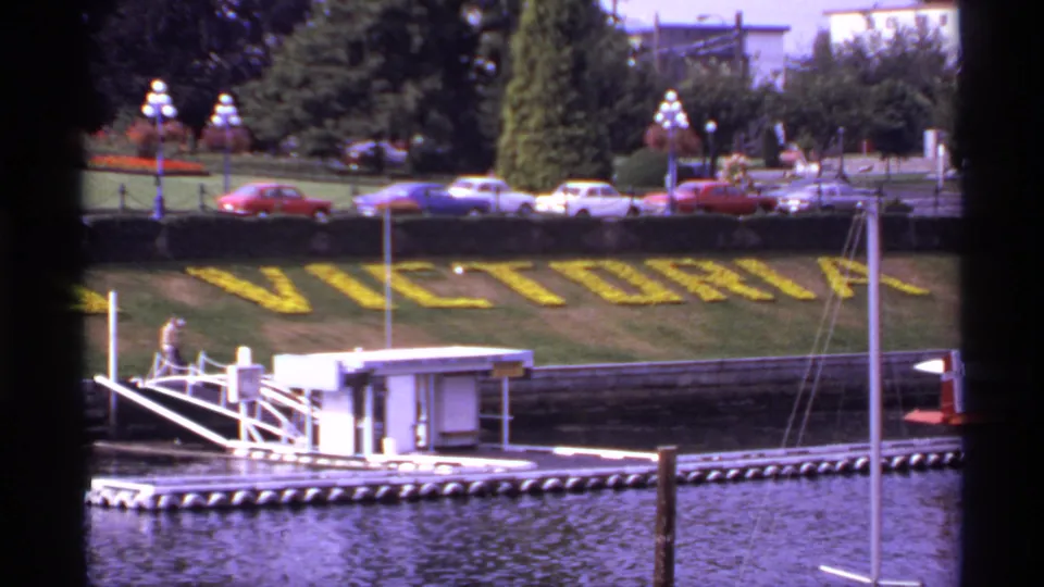 The Victoria floral lettering appears above a harbor dock, with parked cars, trees, lamp posts, boats, and water in a 1974 Canadian travel scene.