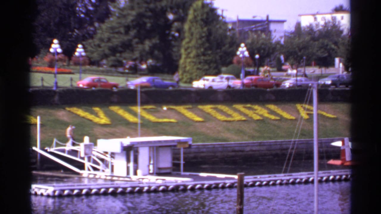 1974 Victoria, Canada home-movie frame showing yellow floral VICTORIA lettering, parked cars, harbor water, and a small dock.