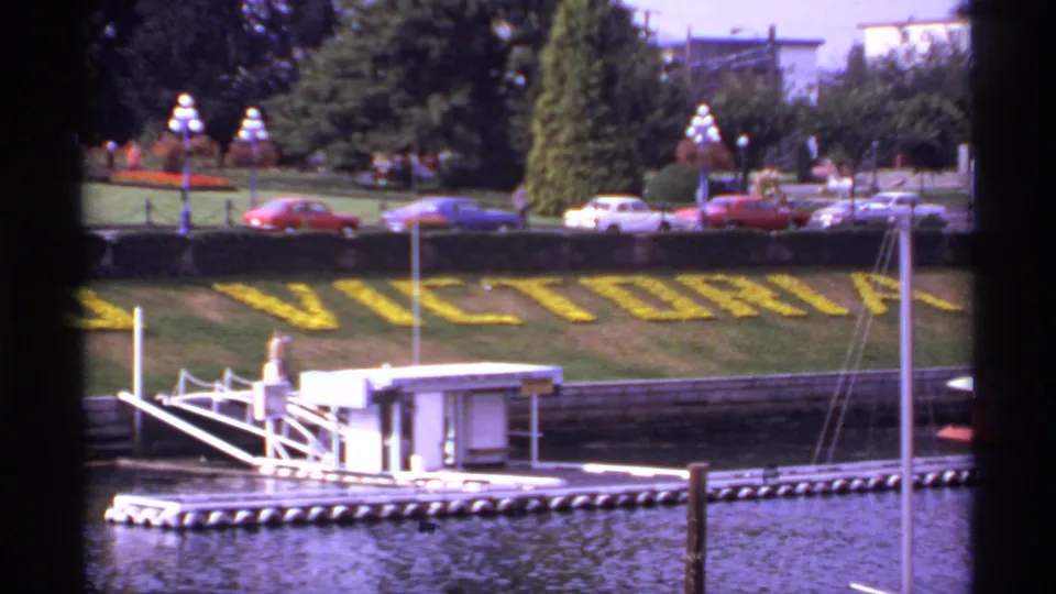 1974 Victoria waterfront view with the yellow VICTORIA floral display, classic cars, trees, lampposts, and dock structures.