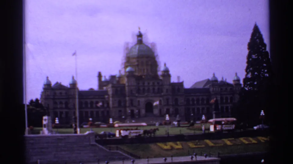 1974 home-movie view of the British Columbia Parliament Buildings across the Victoria waterfront, with a domed roof and people on the lawn.