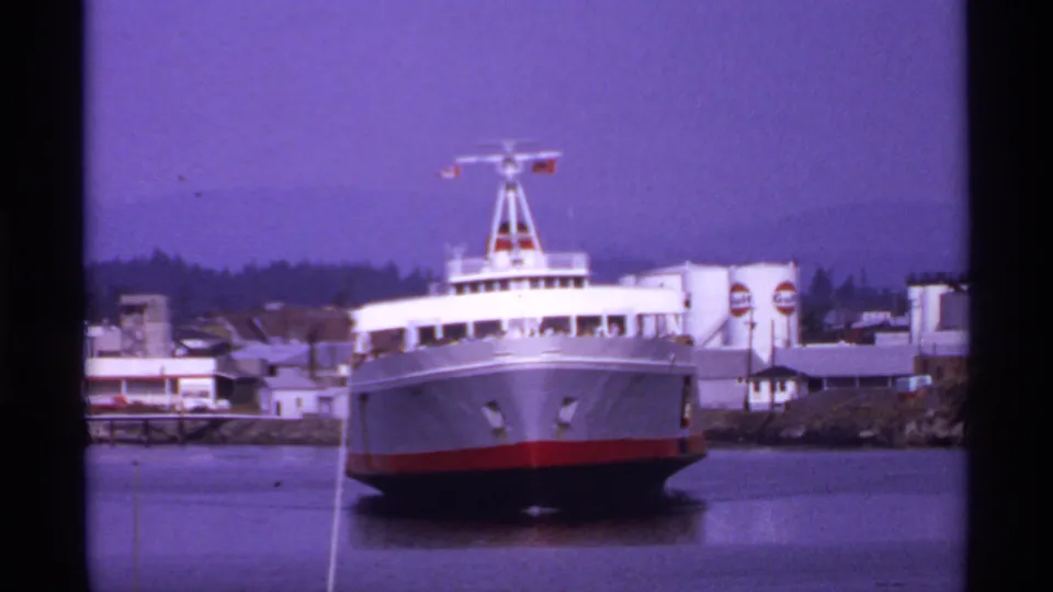 1974 Canadian coastal port view of a large white and red passenger ferry docked near low industrial buildings and fuel signage.