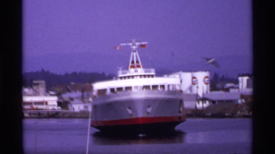 Large ferry bow viewed from shore in a 1974 Canada home movie, with harbor water, dock facilities, and Gulf signs visible behind.