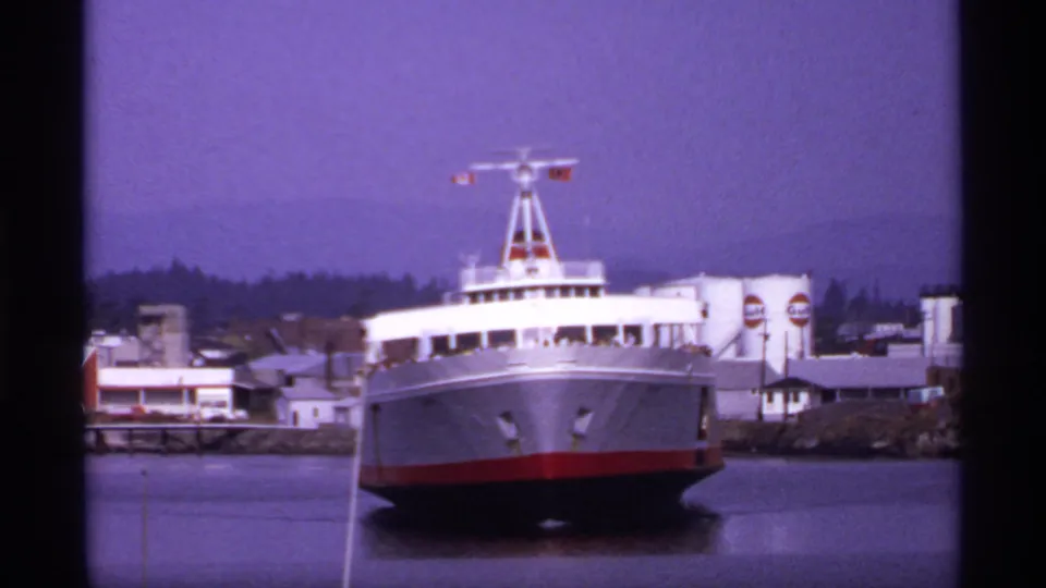 1974 Canadian ferry-port frame showing a white and red passenger ferry at dock, with low buildings, signage, and wooded hills beyond.