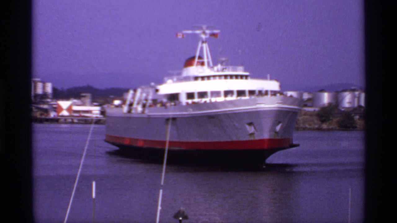 Large white ferry with red lower hull approaching across calm harbor water.