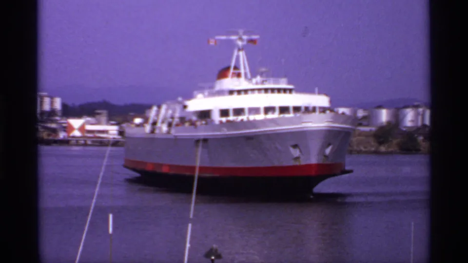 1974 Canadian 8mm frame showing a large passenger ferry nearing shore, with industrial tanks and waterfront structures behind it.