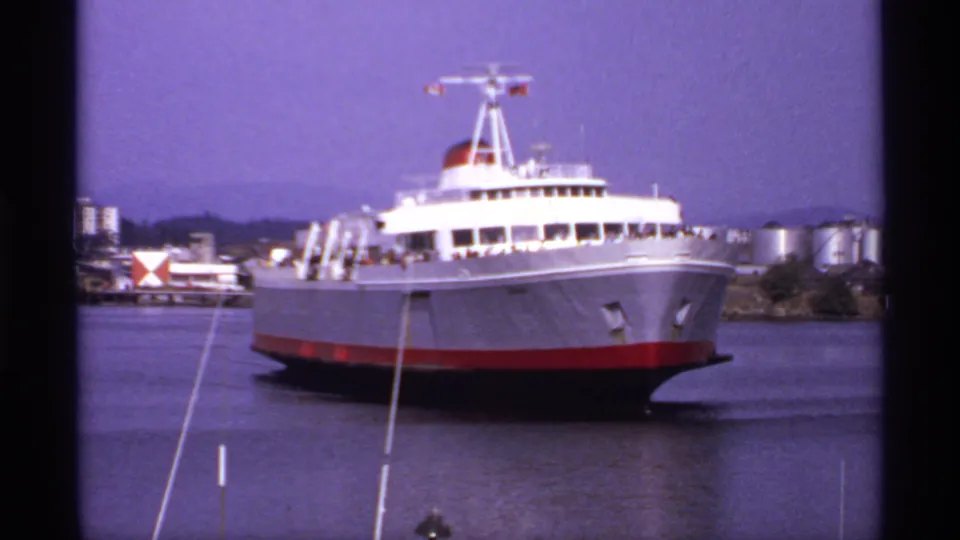 A 1970s Canadian home-movie view of a passenger ferry on calm water, framed by dockside poles and an industrial shoreline.
